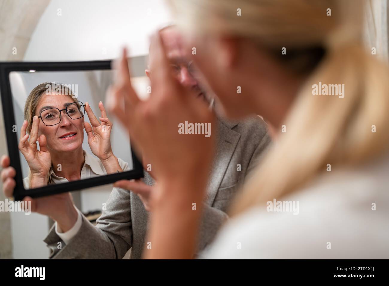 Optician holding a mirror to female customer trying on glasses in a optical store. Stock Photo