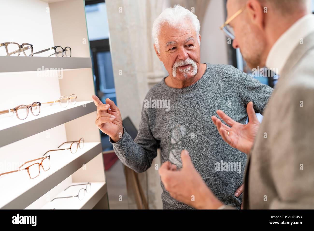 Senior man gesturing during a conversation with an optician in an optical shop with glasses displayed in the background Stock Photo