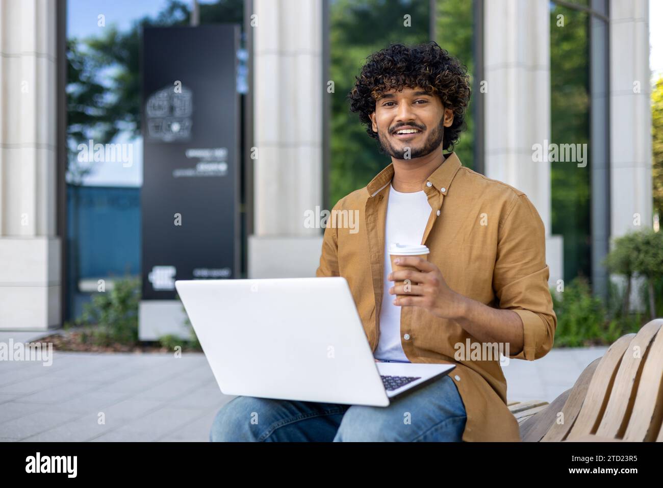 Portrait of a young Indian freelancer man sitting on a bench outside with a laptop, holding a cup of coffee and smiling at the camera. Stock Photo