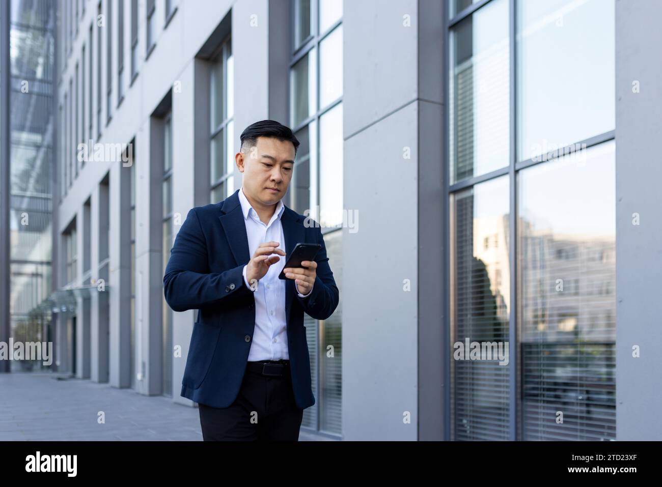 A young Asian man in a business suit is walking down the street near office buildings and using a mobile phone. Stock Photo