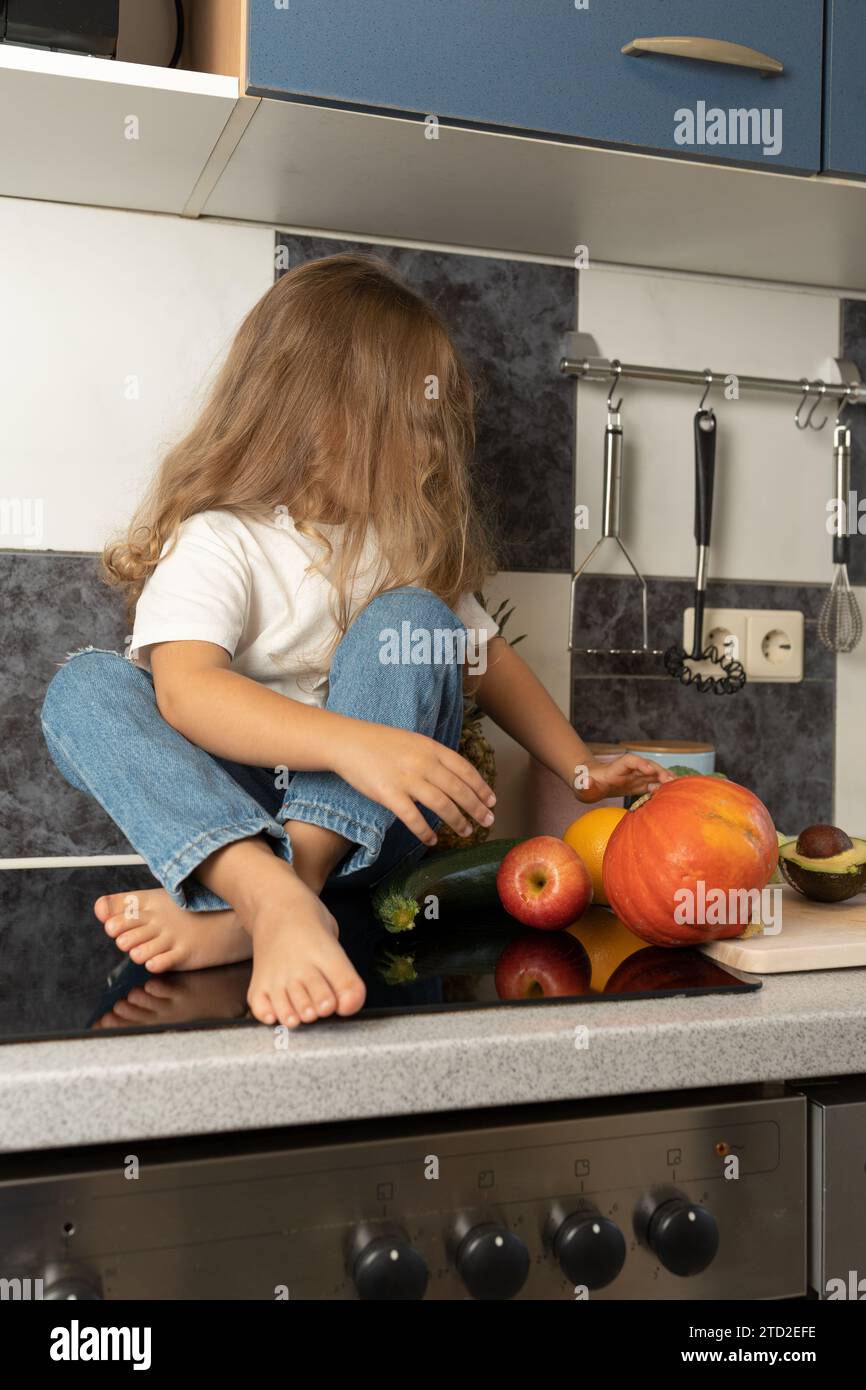 The barefoot child is sitting on the kitchen table. A little girl with long hair. A young woman is cutting fruits and vegetables in the kitchen. Stock Photo