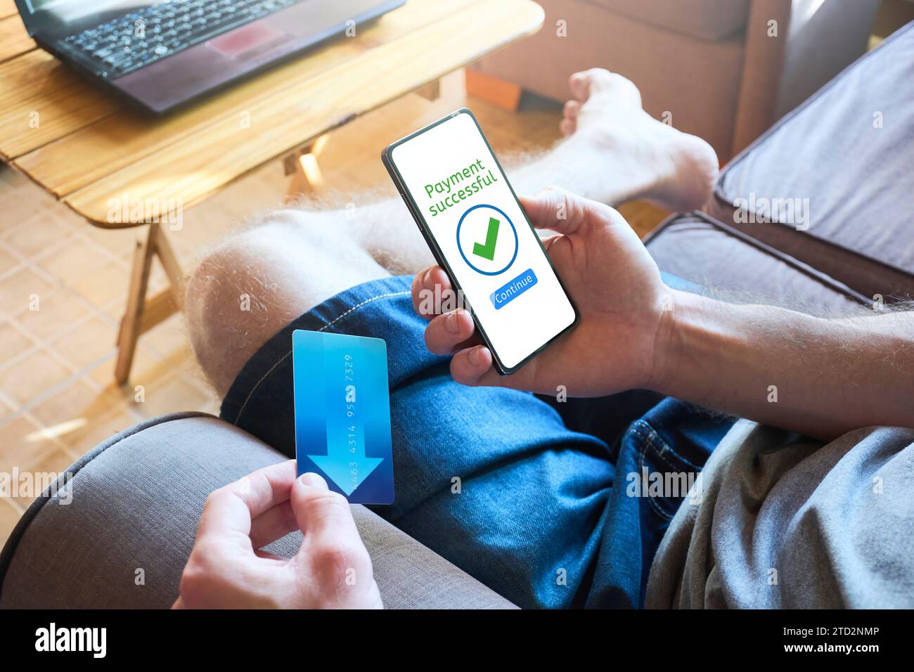 Man using mobile phone for payment while sitting on the couch at home, credit card in hand. Payment successful message on the screen, transaction comp Stock Photo