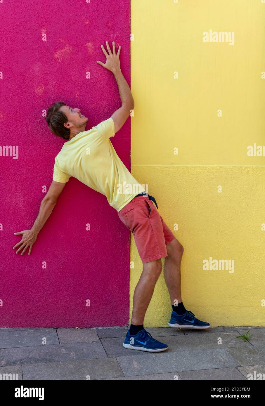 Young man in front of a house wall, with trousers and T-shirt in the same colours as the wall, red and yellow house facade, colourful houses on the Stock Photo