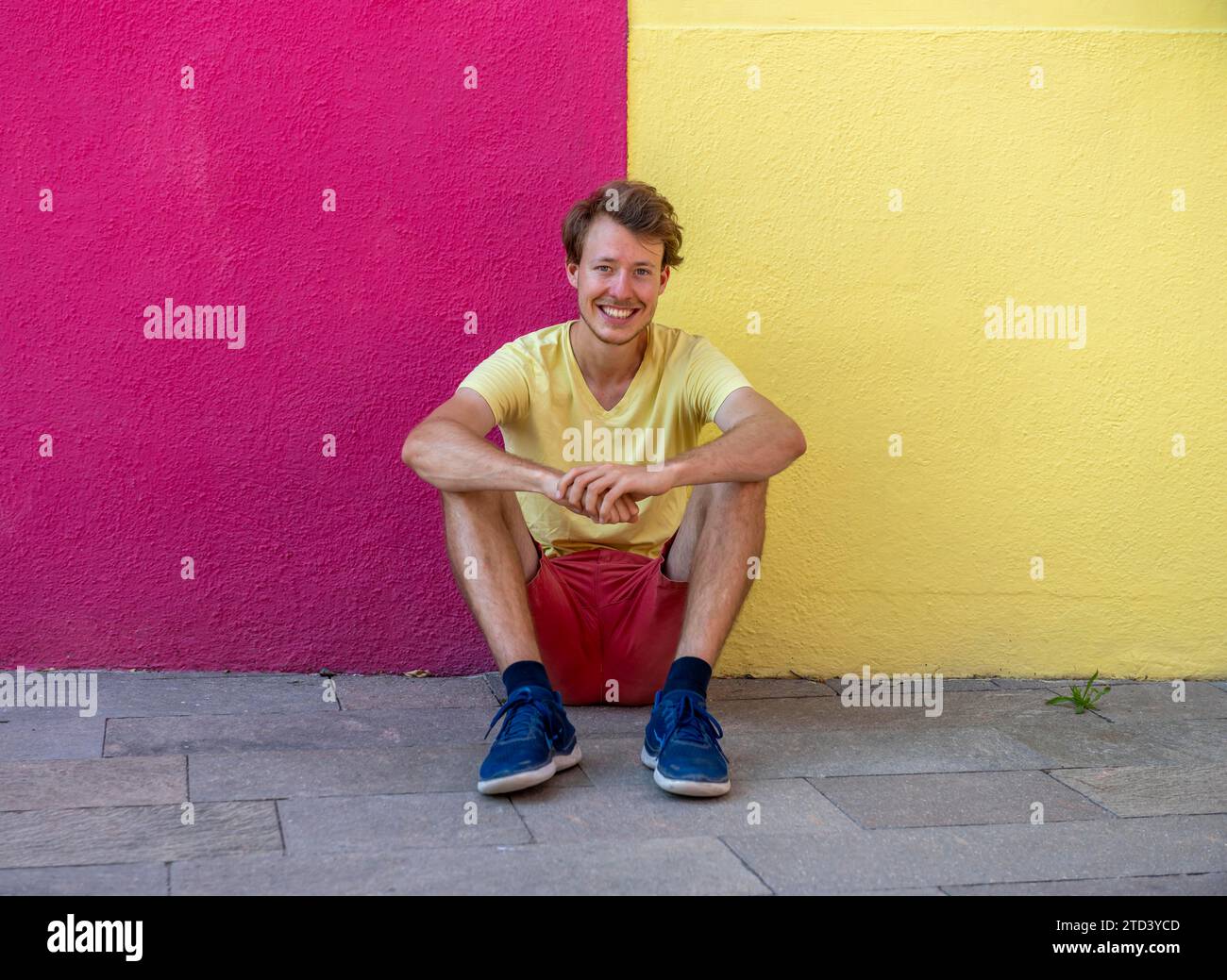 Young man sitting in front of a house wall, with trousers and T-shirt in the same colours as the wall, red and yellow house facade, colourful houses Stock Photo