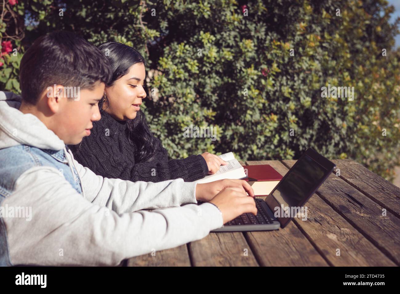 Two children studying with a laptop and books on a wooden table outdoors on a sunny day Stock Photo
