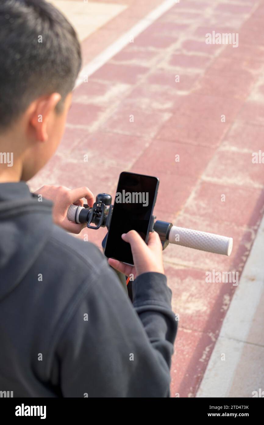 A boy is looking at a smartphone while standing with a scooter on a pathway Stock Photo