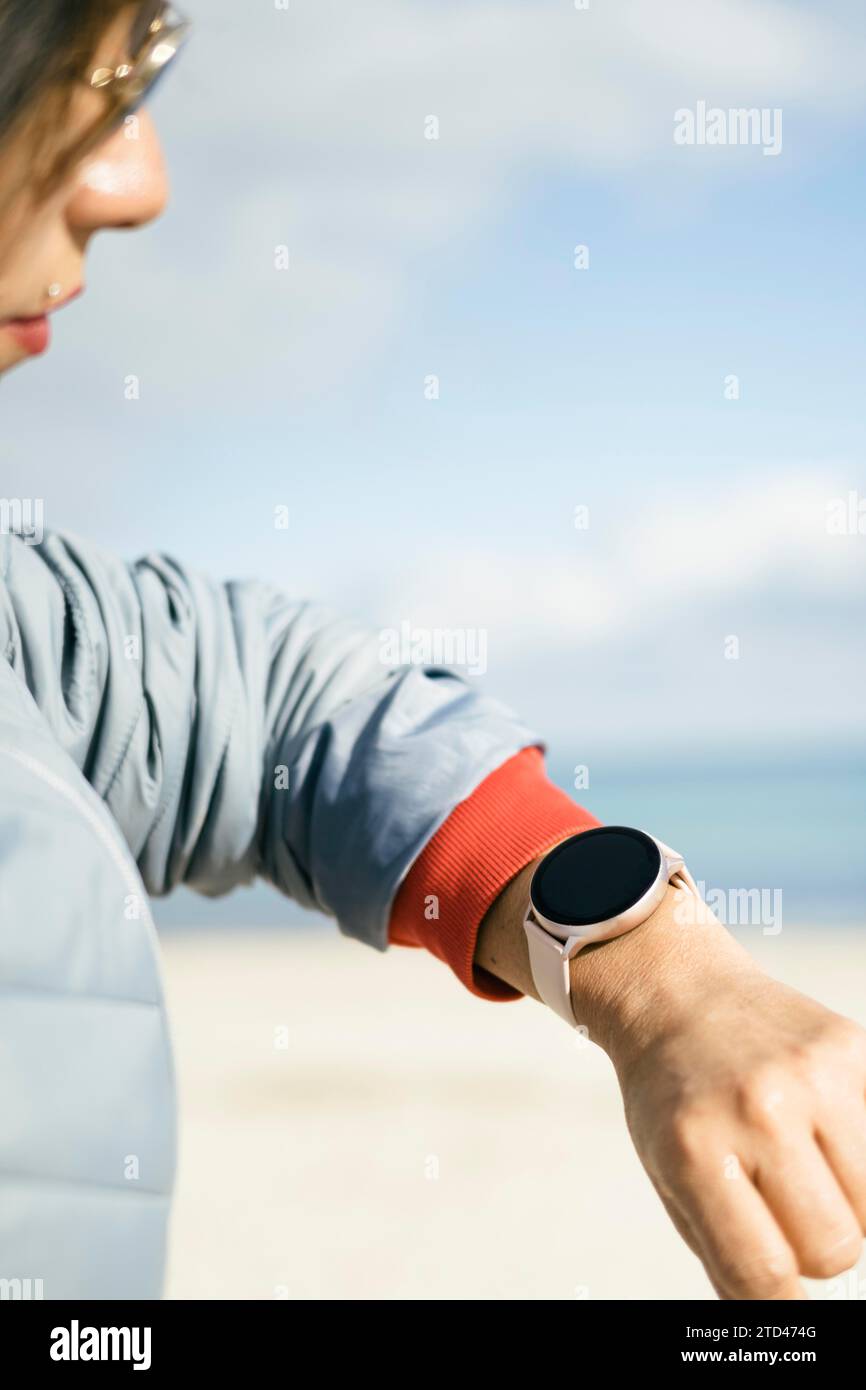 Close-up view of latina woman looking at smart watch, wearing sunglasses blue jacket on a sunny day with clear background of a beach and blue sky to Stock Photo