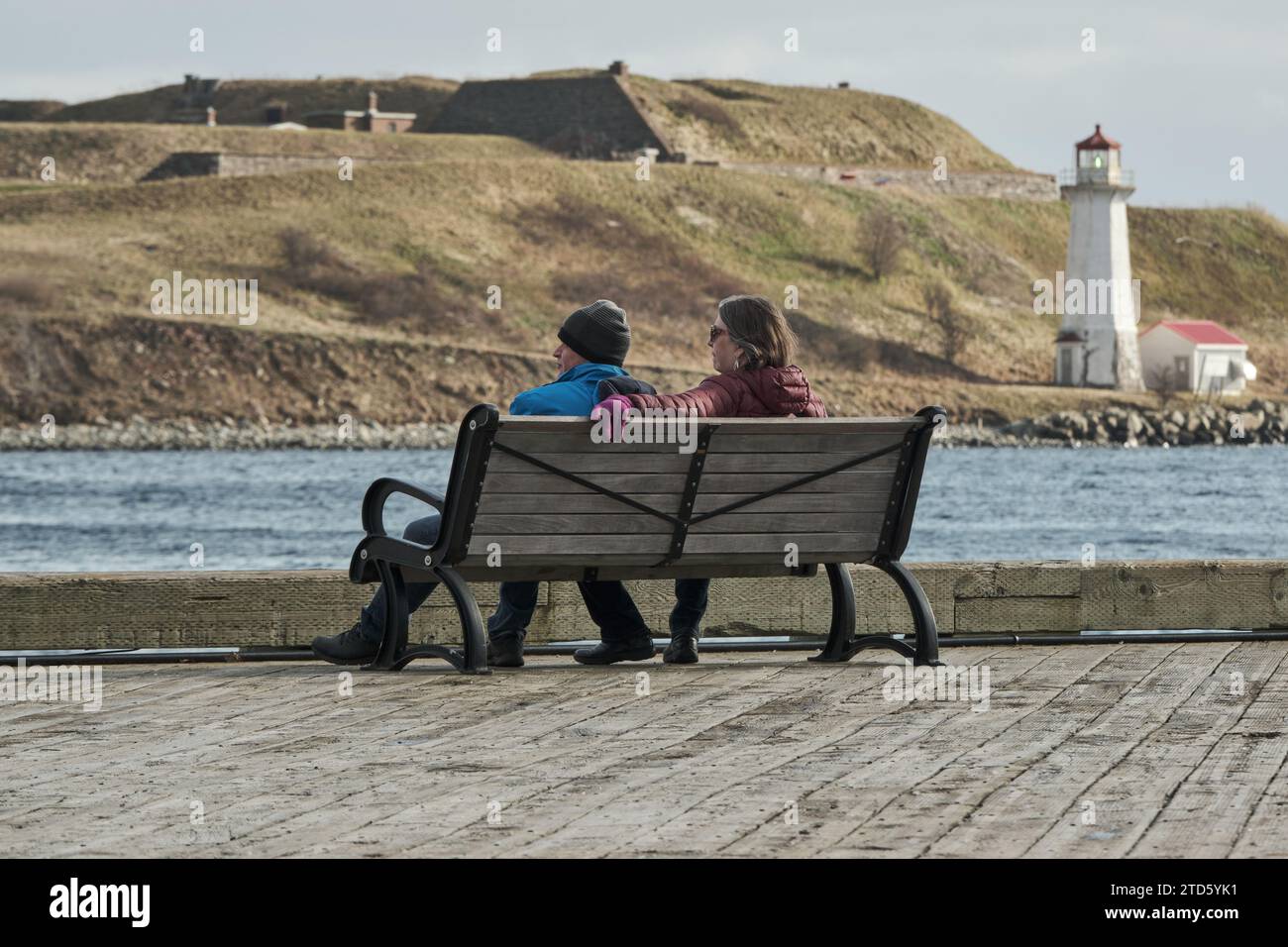 Couple sitting on bench on Halifax waterfront boardwalk, with George Island lighthouse in background Stock Photo