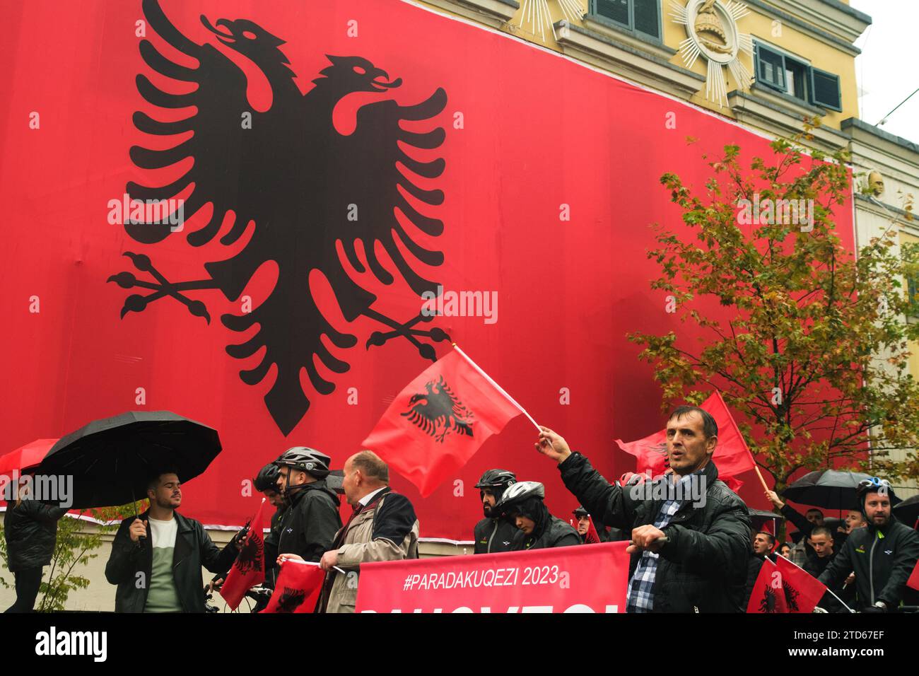 Tirana, Albania - November 28, 2023: On a rainy Independence Day, Tirana City Hall is covered with a massive Albanian flag. Citizens pass by the flag- Stock Photo