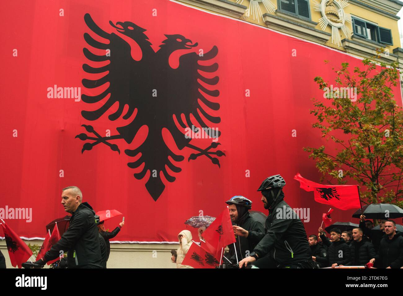 Tirana, Albania - November 28, 2023: On a rainy Independence Day, Tirana City Hall is covered with a massive Albanian flag. Citizens pass by the flag- Stock Photo