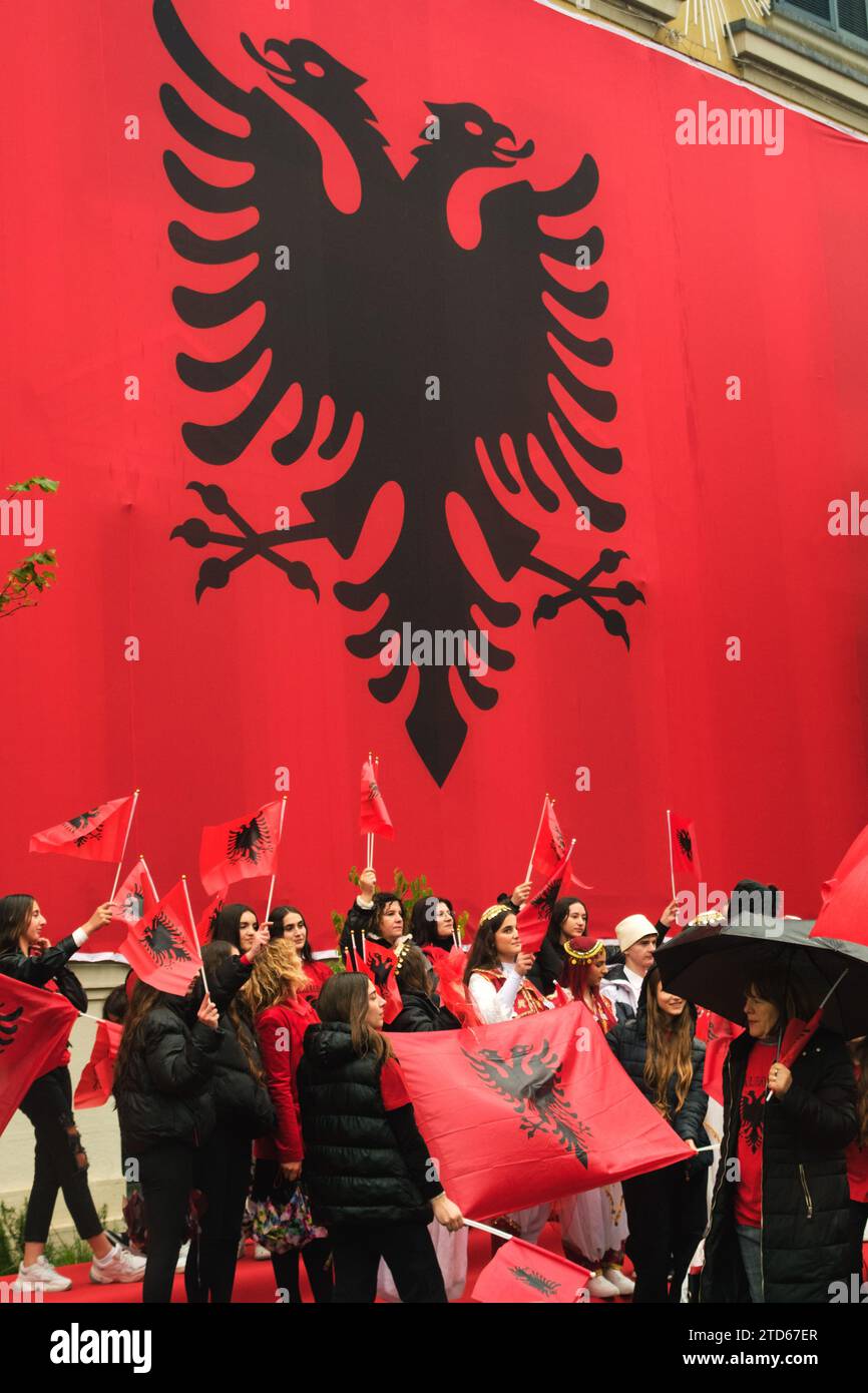 Tirana, Albania - November 28, 2023: On a rainy Independence Day, Tirana City Hall is covered with a massive Albanian flag. Citizens pass by the flag- Stock Photo