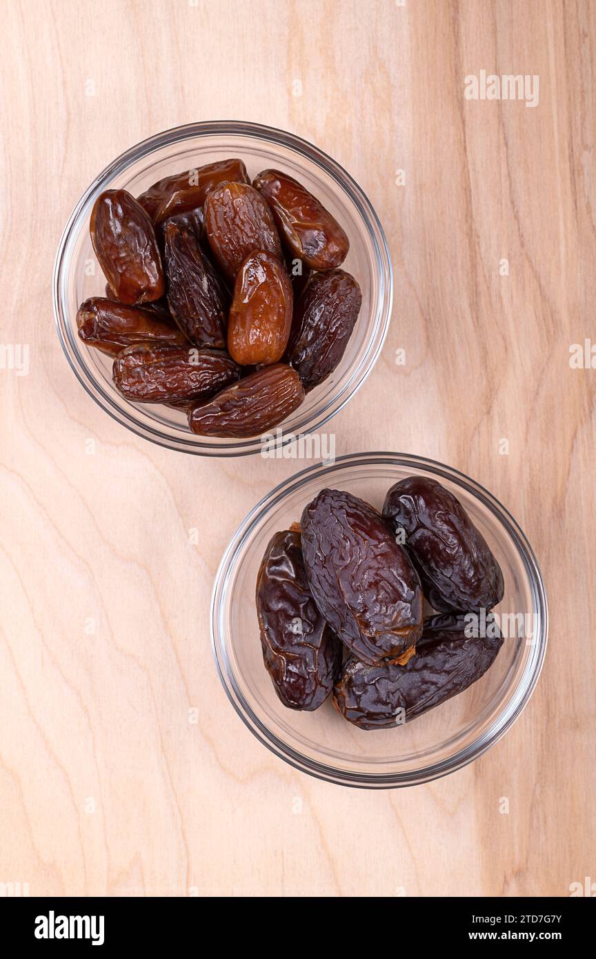 Deglet Nour and Medjool dates, in glass bowls, on a wooden panel, from above. Sun-dried and sweet cultivars of dates. Stock Photo