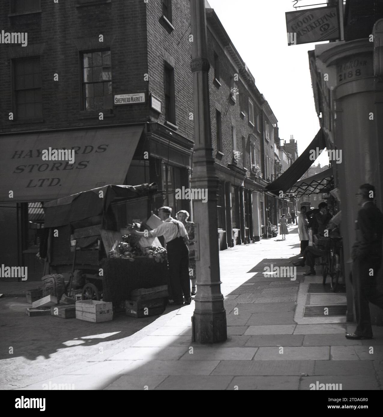 1950s, historical, a female shopper with a male trader at market stall, Shepherd Market, in Mayfair, London, England, UK. On the corner of the paved alleyway, Hayward Stores and a sign for Good Housing Keeping magazine at 1'9 a month. Located in Mayfair off Piccadilly, Shepherd Market is a charming part of central London with its small square and narrow side streets, being known as a 'village in Piccadilly'. Stock Photo