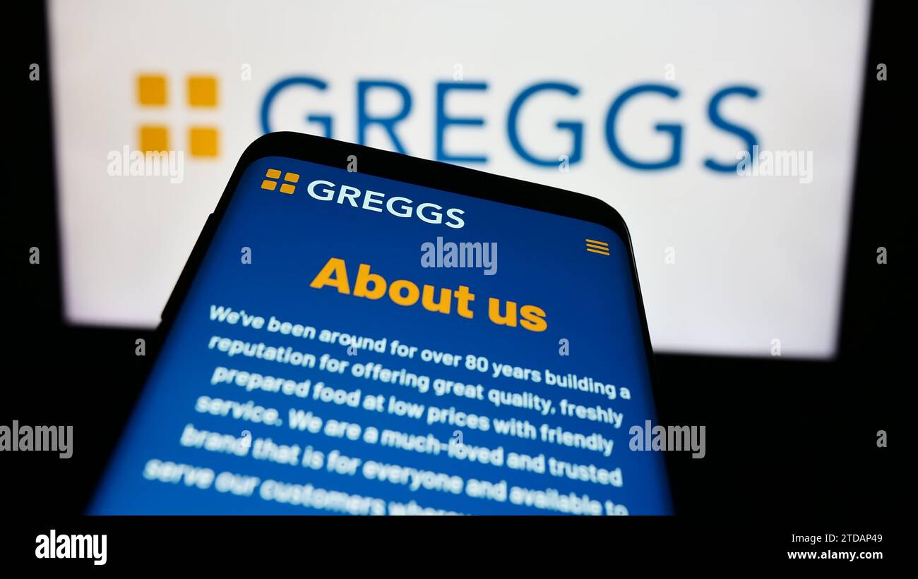 Mobile phone with website of British bakery chain company Greggs plc in front of business logo. Focus on top-left of phone display. Stock Photo