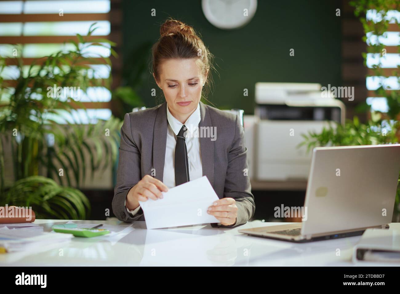 New job. pensive modern 40 years old woman employee in modern green office in grey business suit with laptop opening letter. Stock Photo