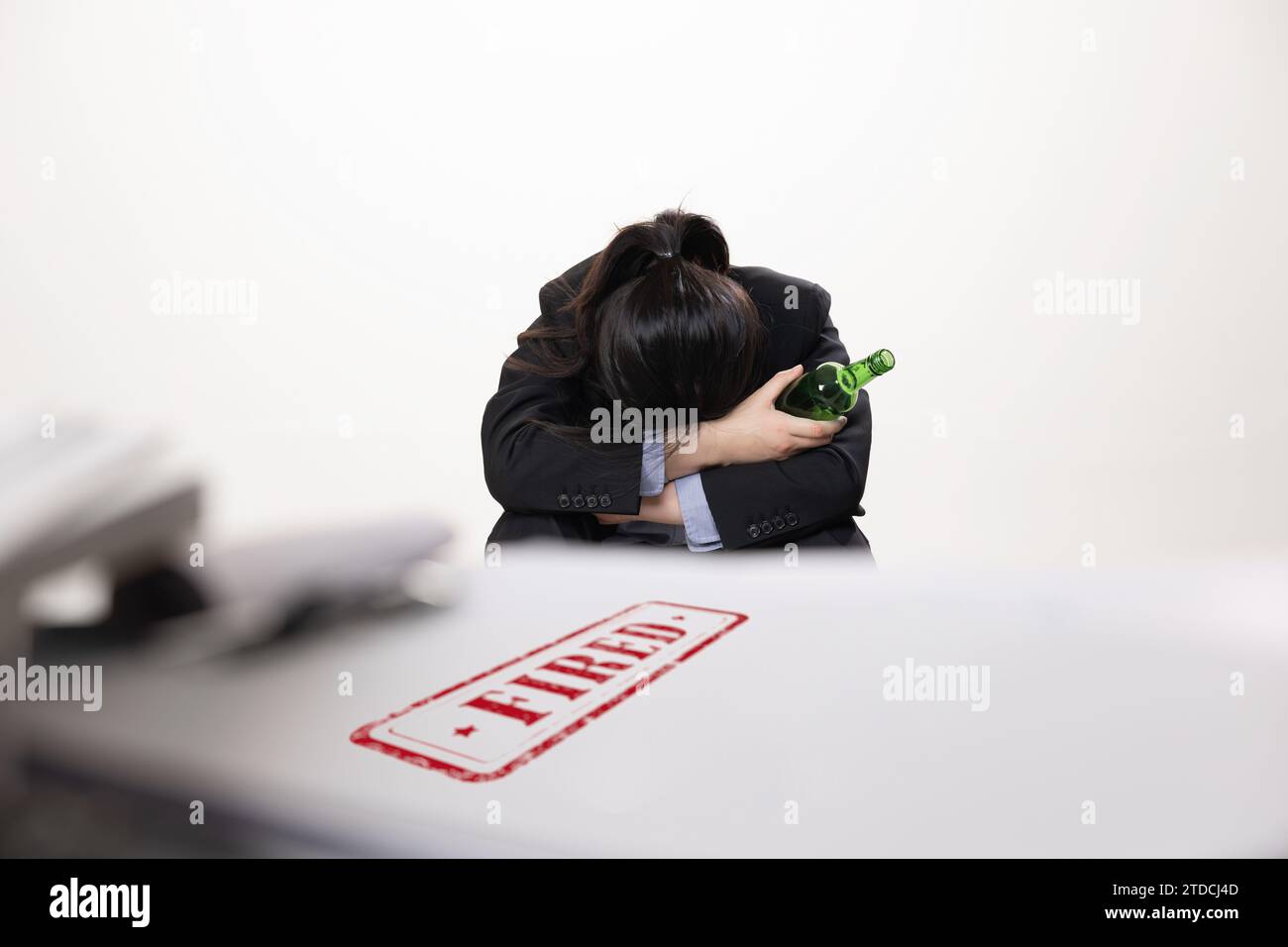 A female office worker in despair holding a dismissal notice and a bottle of alcohol on her desk Stock Photo