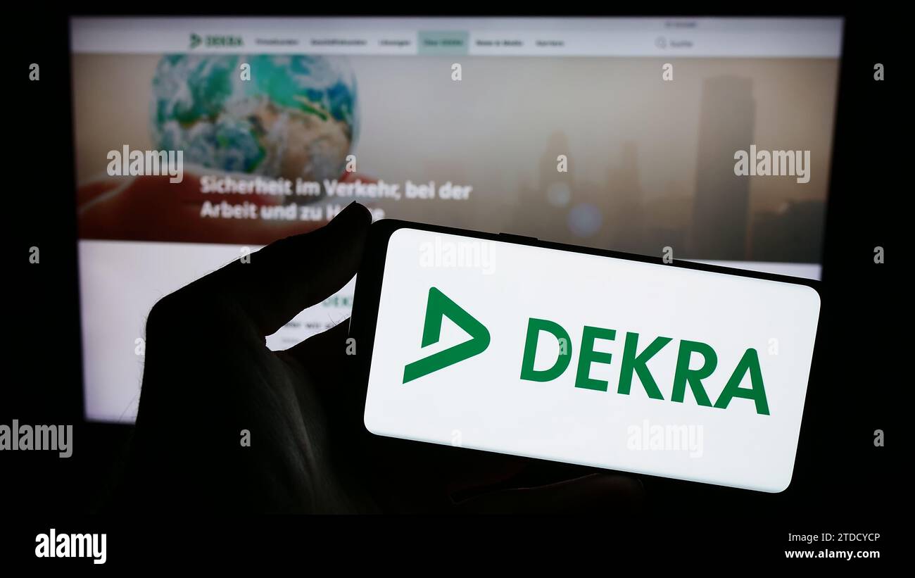 Person holding smartphone with logo of German expert organisation DEKRA in front of website. Focus on phone display. Stock Photo