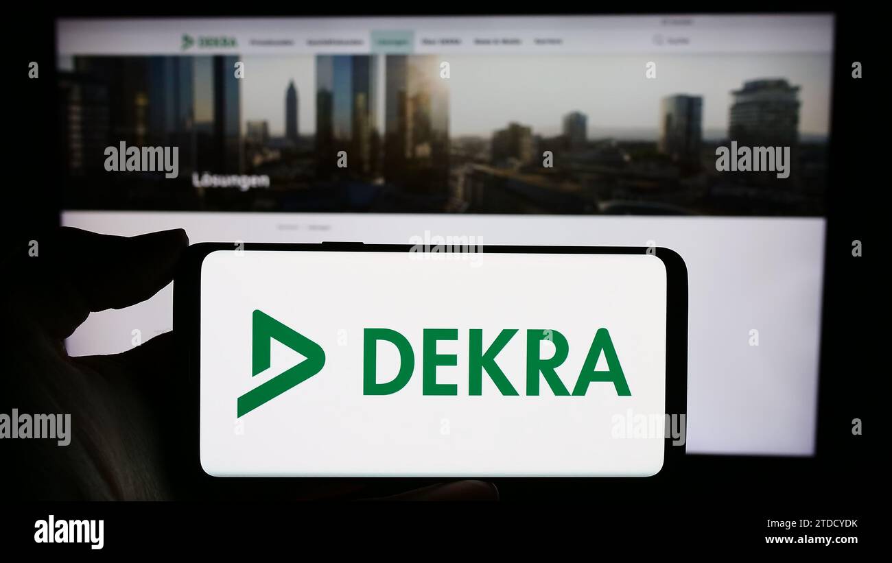 Person holding cellphone with logo of German expert organisation DEKRA in front of business webpage. Focus on phone display. Stock Photo