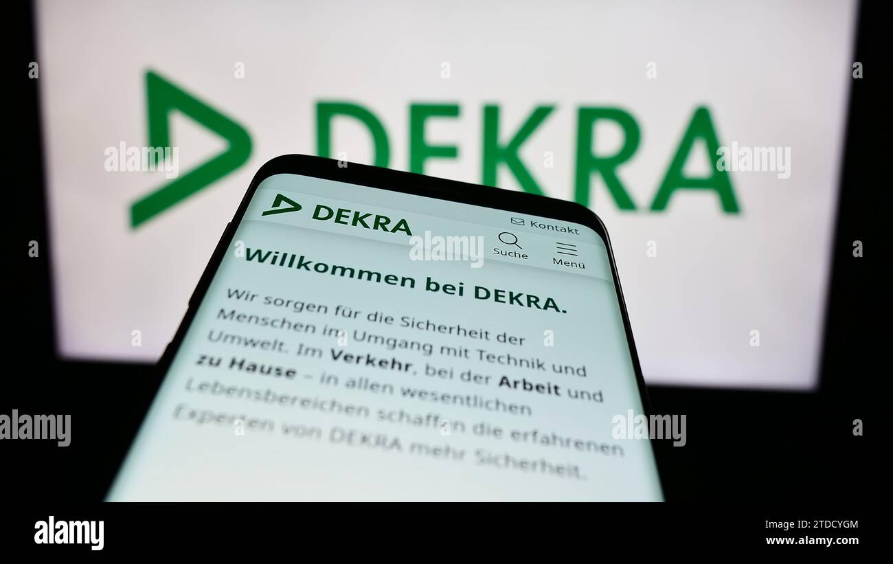 Mobile phone with website of German expert organisation DEKRA in front of company logo. Focus on top-left of phone display. Stock Photo