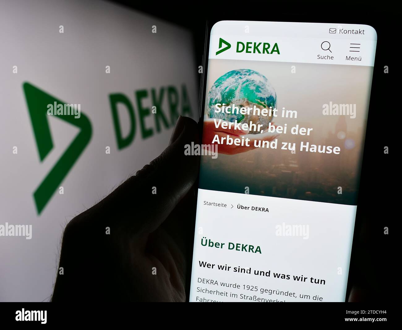 Person holding cellphone with webpage of German expert organisation DEKRA in front of business logo. Focus on center of phone display. Stock Photo