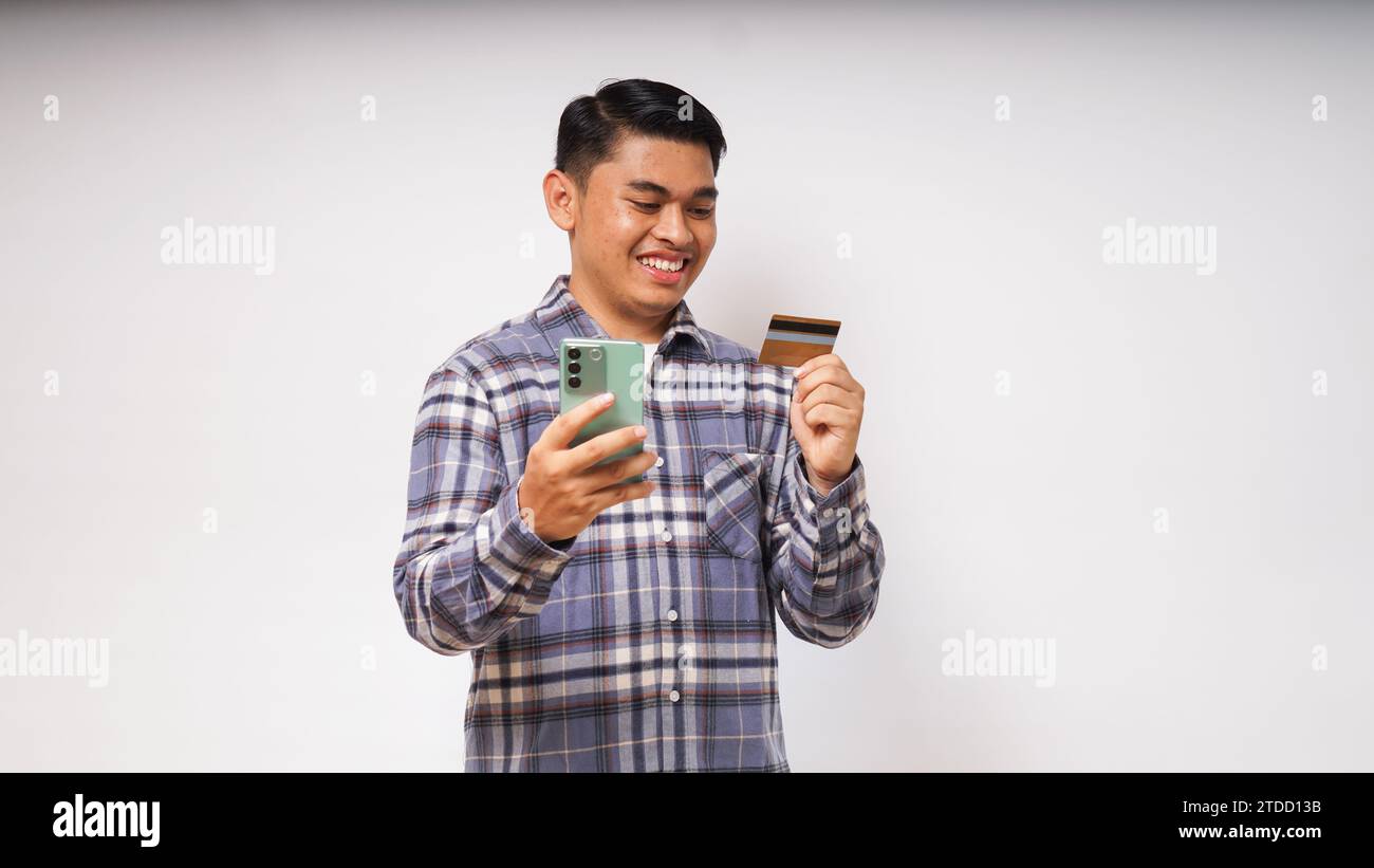Portrait of satisfied happy Asian young man holding smart phone and credit card, entering data, online shopping, wearing casual shirt. Indoor studio s Stock Photo