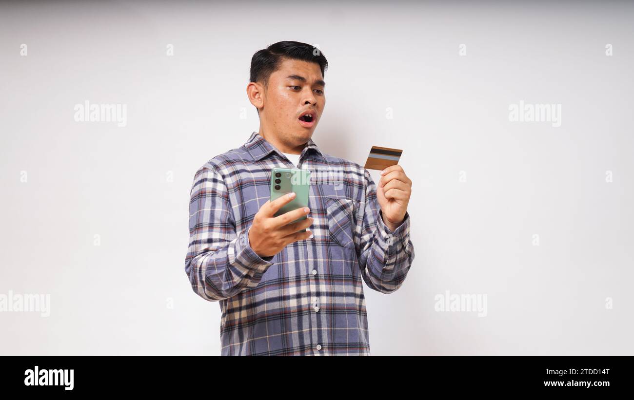 Asian young man with Shocked expression holding credit card and smartphone, looking at the smartphone screen with big eyes, Indoor studio shot isolate Stock Photo