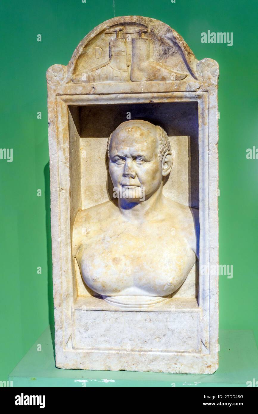 Funerary stele of Caius Julius Helius. Last 20 years of the 1st century AD. Lunense marble - The shoemaker Caius Julius Helius owned a tomb near the ancient Via Triumphalis, where this funerary stele was discovered. Thanks to the dedication on the base, we know that his shop was located at Porta Fortinalis, near the Capitoline Hille. Judging by the two shoe shapes depicted in the upper area of the aedicule, the shoemaker must have specialised in making lace-up sandals, which were mostly worn by soldiers. The figure is depicted proudly: the features of his face are realistic and very expressive Stock Photo