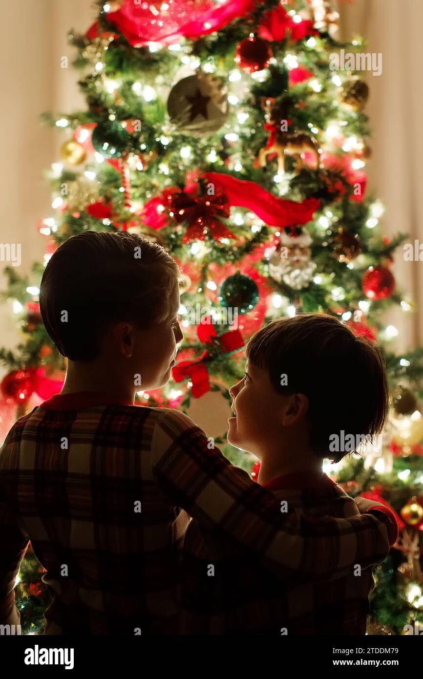 two brothers in pajamas hugging in front of a lit up christmas tree Stock Photo