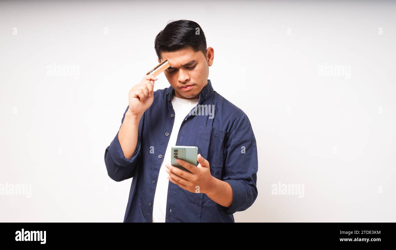 Asian young man with Shocked expression holding credit card and smartphone, looking at the smartphone screen with big eyes, Indoor studio shot isolate Stock Photo