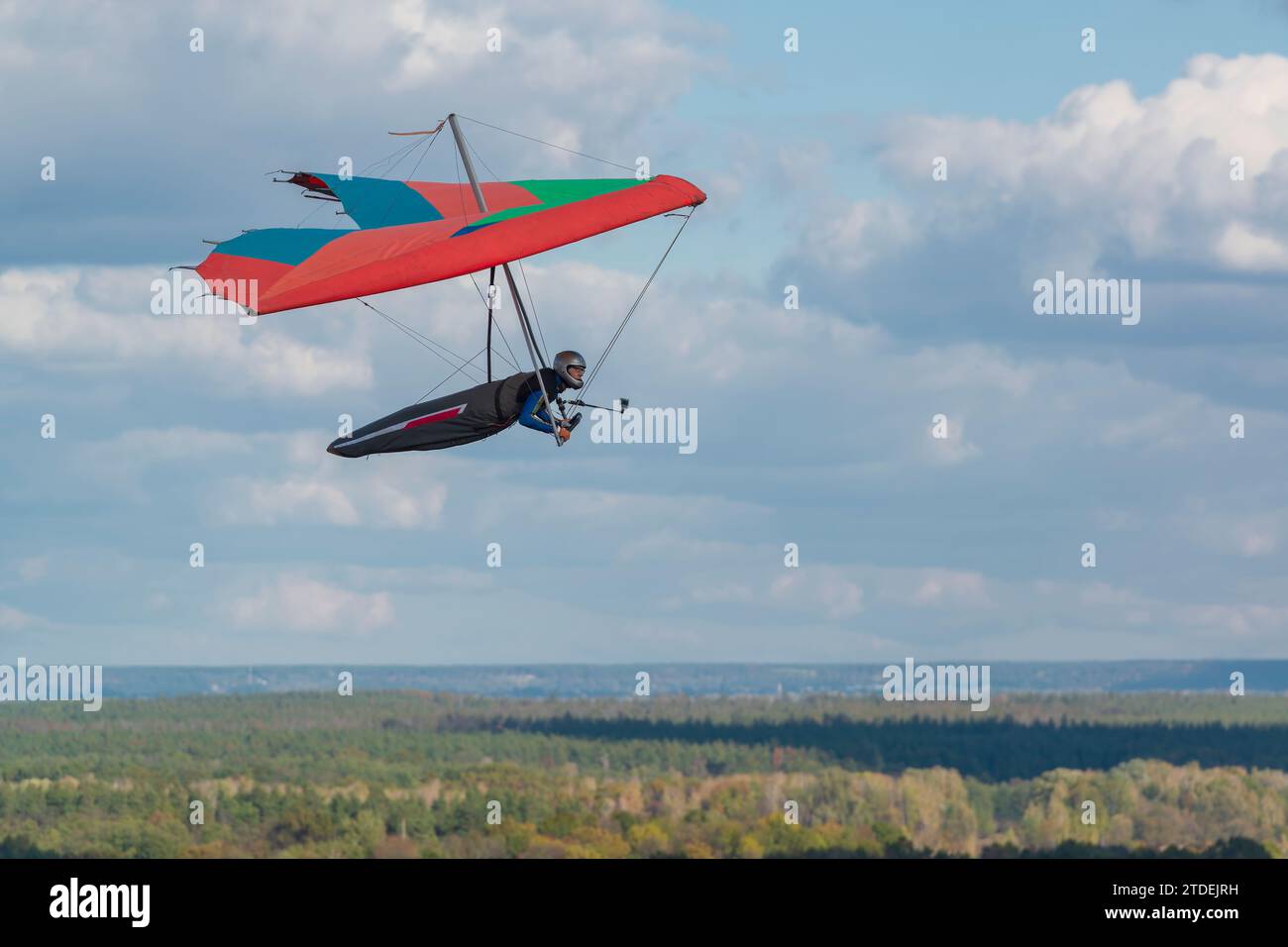 Colorful red hang glider wing soars over fields and forests with blue sky and clouds on the background. Stock Photo