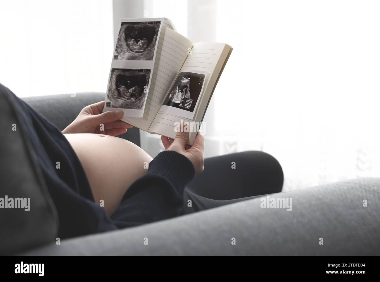 A pregnant woman is sitting on a sofa by the window in the living room and looking at an ultrasound picture of her fetus Stock Photo