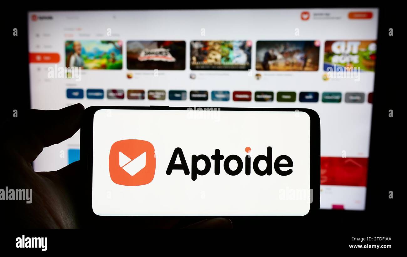 Person holding cellphone with logo of Portuguese app marketplace company Aptoide S.A. in front of business webpage. Focus on phone display. Stock Photo