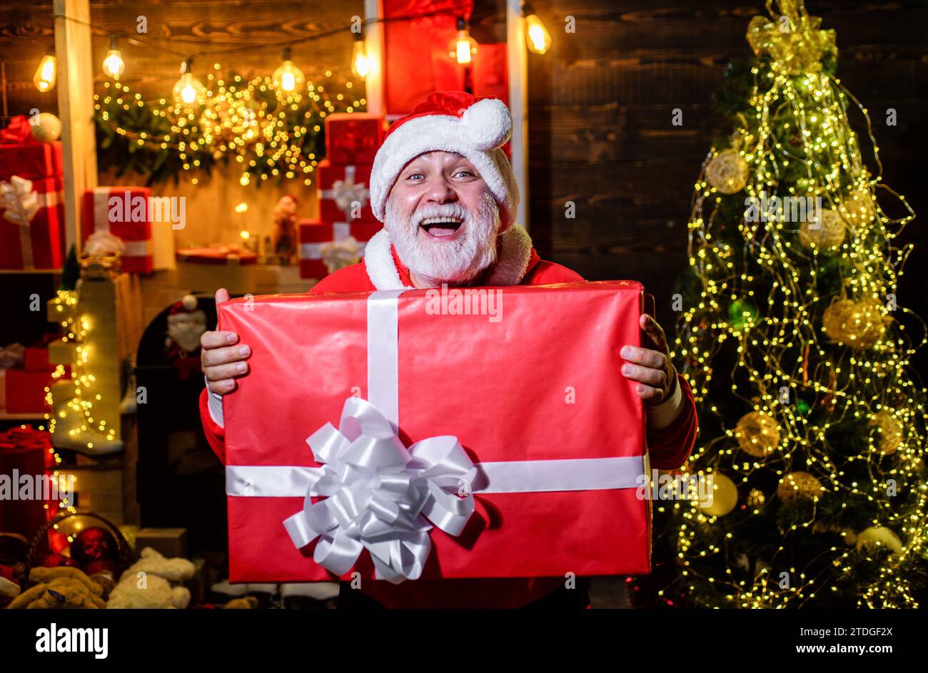 Christmastime. Smiling Santa Claus with Christmas gift box in room decorated for Christmas. Winter family holiday. Bearded man in Santa hat with Stock Photo