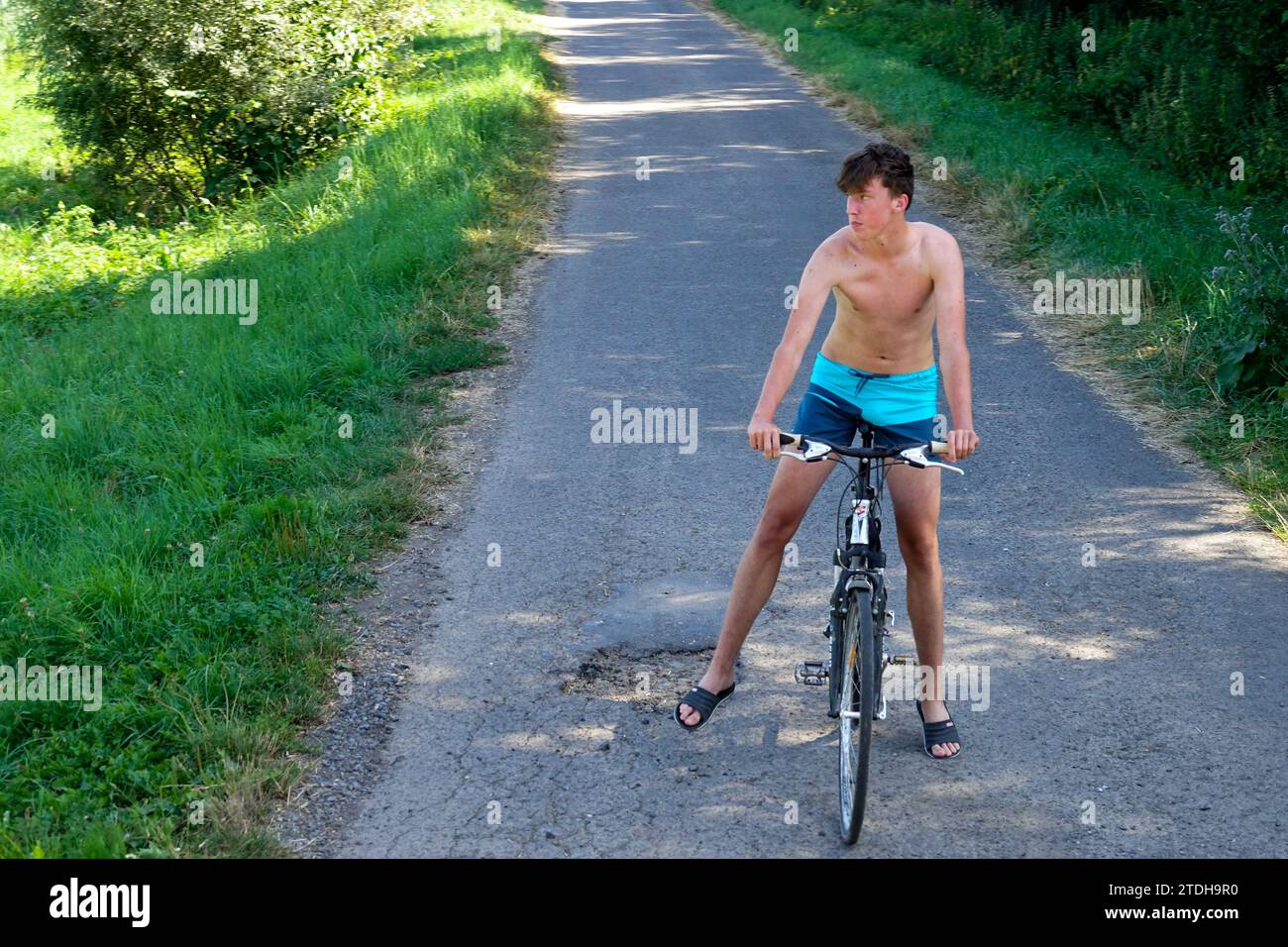 young man alone 20s on a bicycle on a rural road, summer, only in shorts riding bike without helmet, hot weather Stock Photo