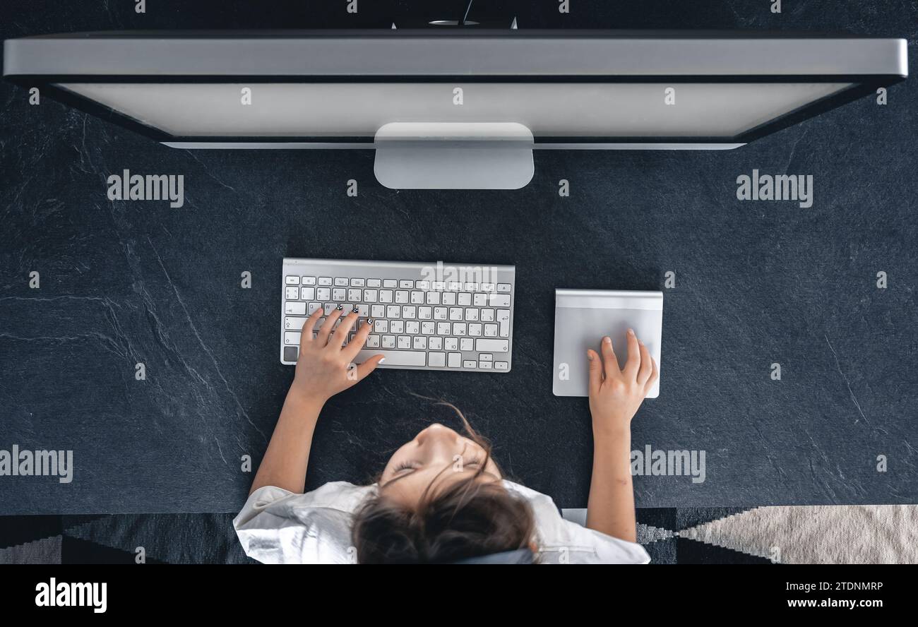 A little girl sits in front of a computer screen at a table, top view ...