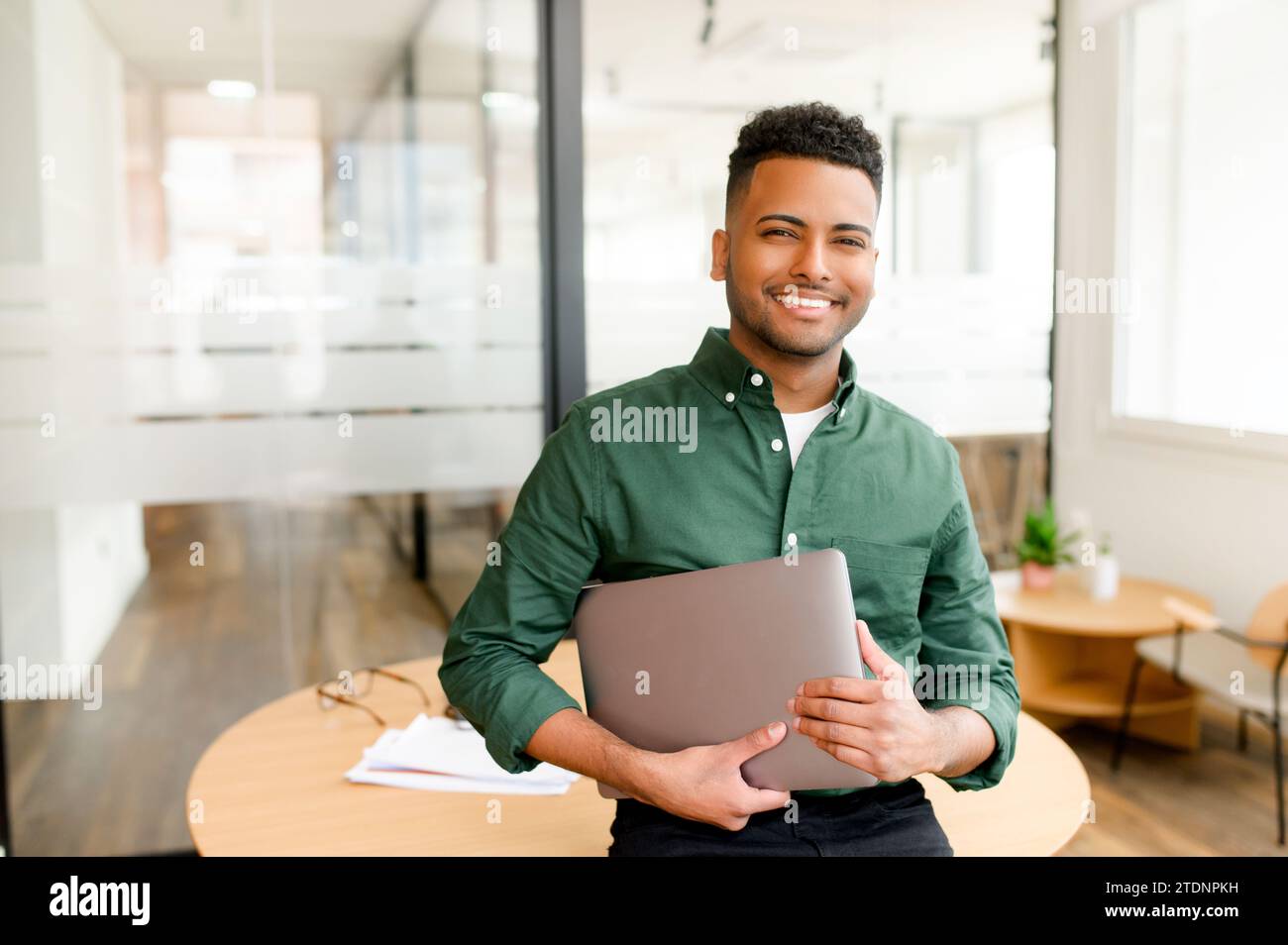 Portrait of confident indian businessman standing with laptop, male office employee in smart casual wear looking at the camera and smiles, leaned back at te desk, ceo, manager, sturtup owner Stock Photo
