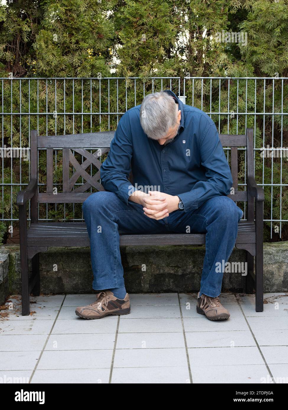 Man with grey hair and full beard, 50-59 years old, sitting on a bench on the terrace outside, hanging his head, thoughtful, sad Stock Photo