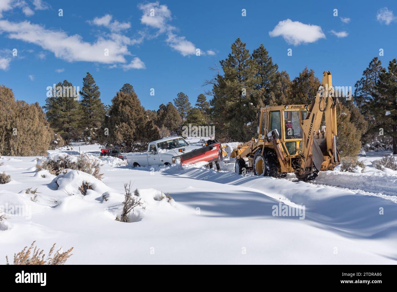 White pickup truck stuck in the snow and a yellow Ford backhoe on a remote road in the Southern Rocky Mountains in Northern New Mexico, United States. Stock Photo