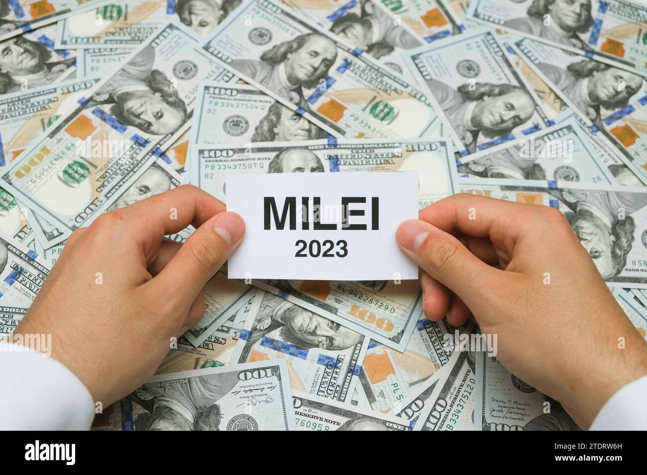 Anonymous Male Hands Holding A Little Paper With The Word 'Milei 2023' On It. Argentina's New President. $100 Dollar Bills Background Stock Photo