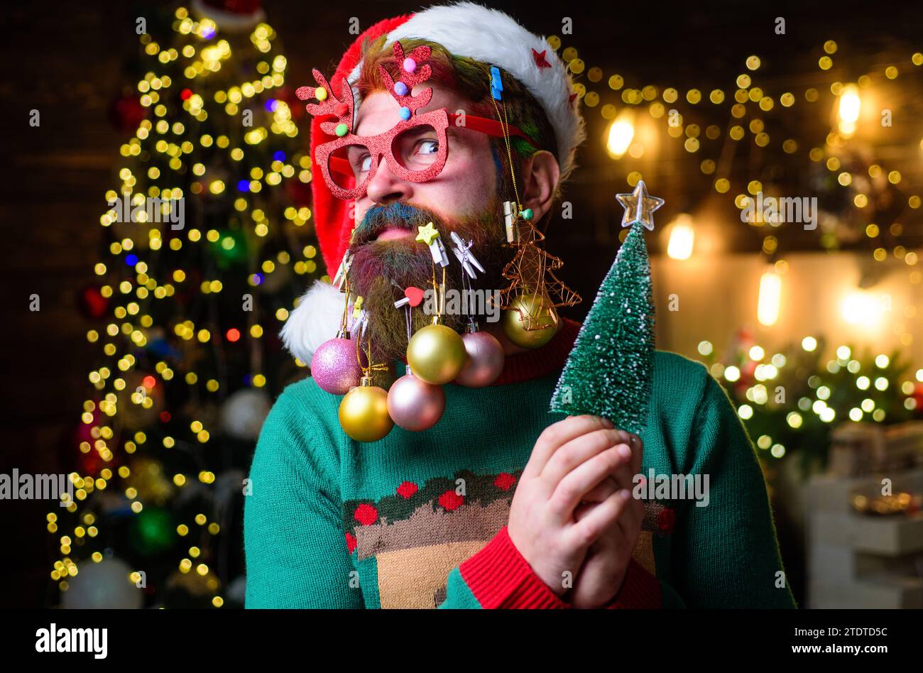 Christmas barbershop. Surprised man with New Year decoration balls in beard with fir-tree. Bearded man with dyed hair in Santa hat and party glasses Stock Photo