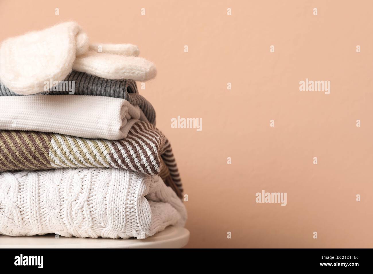 Stack of folded sweaters with gloves on stool against beige background Stock Photo
