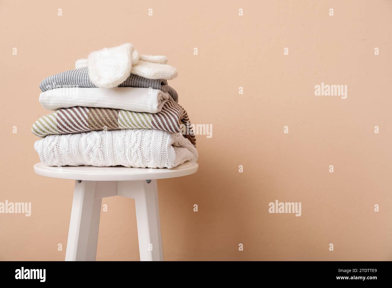 Stack of folded sweaters with gloves on stool against beige background Stock Photo