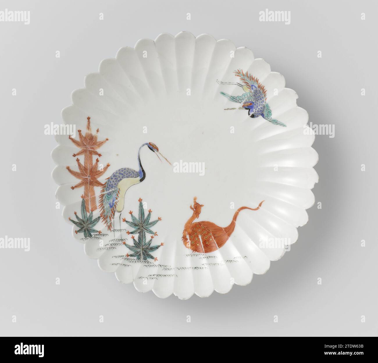 Fluted saucer-dish with cranes, minogame and pine shoots, anonymous, c. 1670 - c. 1690 Scale of porcelain with a ribbed wall and lobed edge, painted on the glaze in blue, red, green, yellow, black and gold. On the flat a standing crane between pine shoots, looking at a minog game on the floor. A second crane in the air. Some chips in the edge. Arita, Kakiemon. Japan porcelain. glaze. gold (metal) painting / gilding / vitrification Scale of porcelain with a ribbed wall and lobed edge, painted on the glaze in blue, red, green, yellow, black and gold. On the flat a standing crane between pine sho Stock Photo