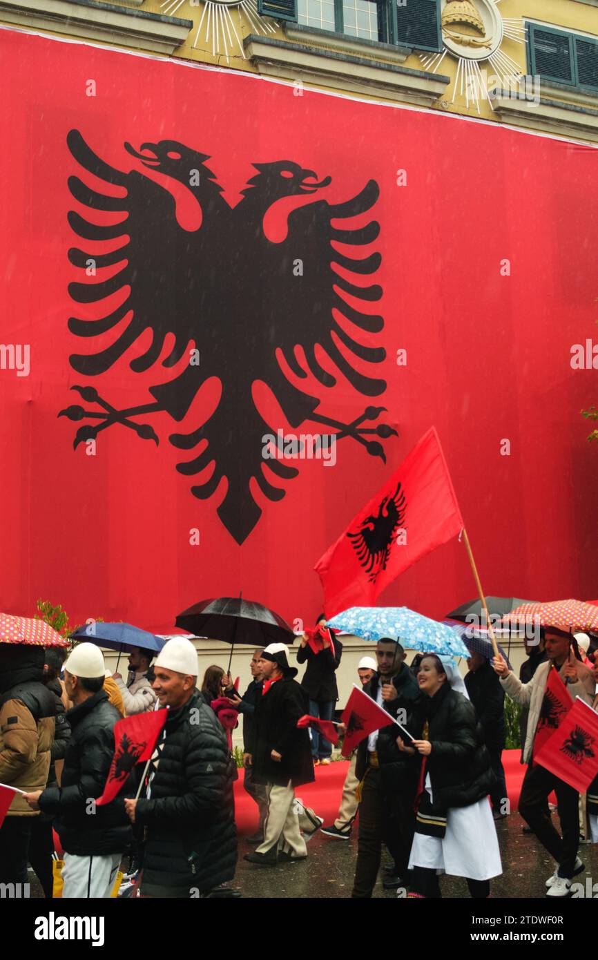 Tirana, Albania - November 28, 2023: On a rainy Independence Day, Tirana City Hall is covered with a massive Albanian flag. Citizens pass by the flag- Stock Photo