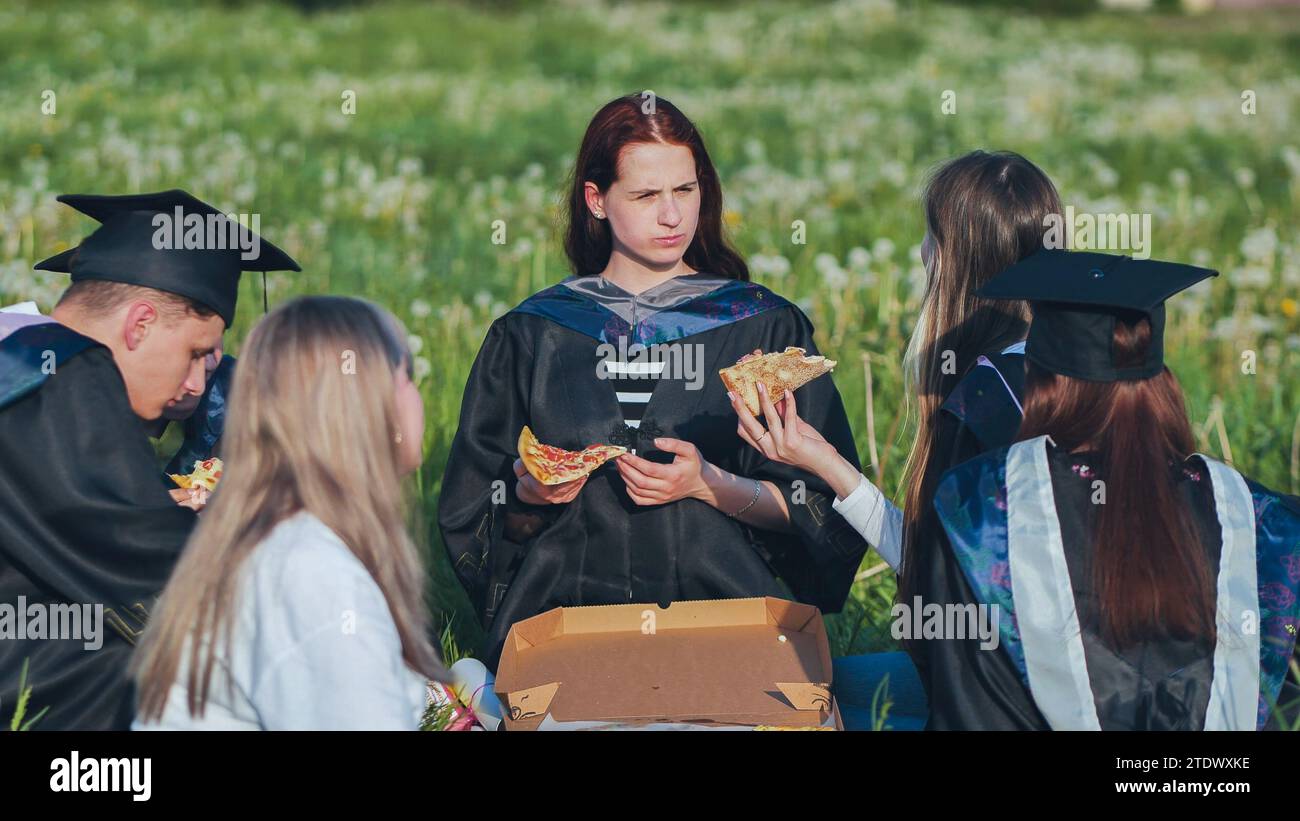 Graduates in black suits eating pizza in a city meadow Stock Photo - Alamy