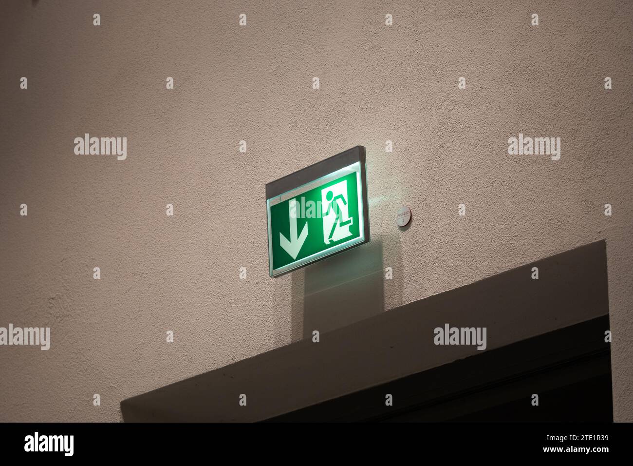 Escape route sign above a door on a wall. The illuminated emergency exit symbol is mandatory in public buildings in Germany. The arrow shows the exit. Stock Photo