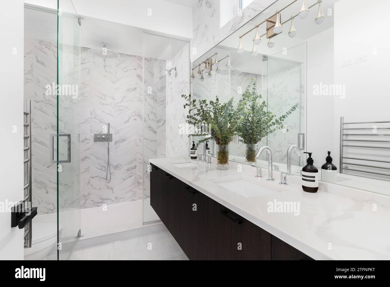 A beautiful bathroom with a floating dark wood cabinet, marble countertop with decorations, and a herringbone tiled walk-in shower. No brands or label Stock Photo