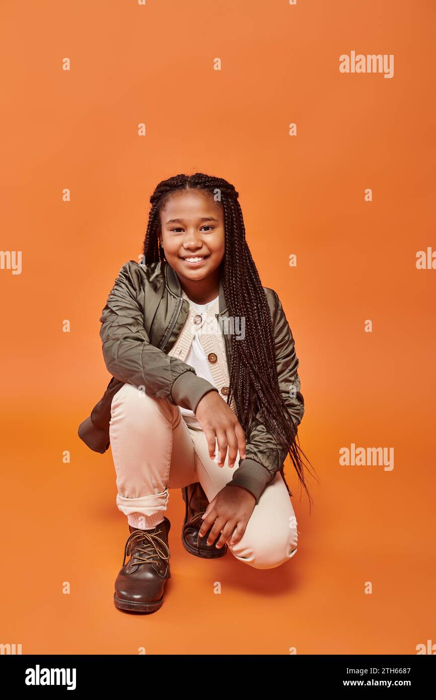 cheerful african american girl with braids in winter jacket squatting and smiling at camera Stock Photo