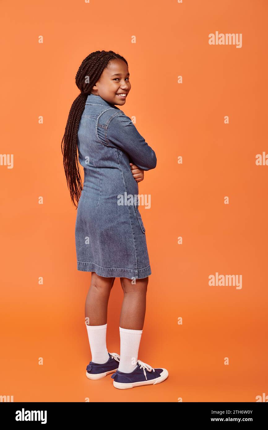 full length of positive african american girl in denim dress standing on orange background Stock Photo