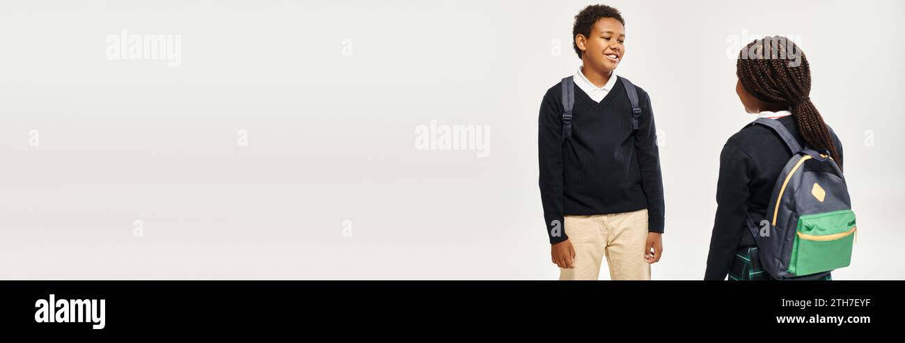 cheerful african american schoolkids with backpacks standing and chatting on grey background, banner Stock Photo